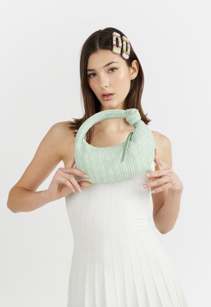 Donna con capelli castani lisci che indossa un vestito bianco e fermagli colorati tiene in mano una borsa verde pallido strutturata con manico annodato.
