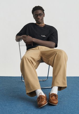 Młody mężczyzna w okularach siedzi zrelaksowany na metalowym krześle, ubrany w czarny t-shirt, beżowe spodnie, białe skarpetki i brązowe skórzane buty na niebieskim dywanie.