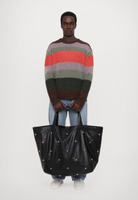 Black leather tote bag with silver studs, large rectangular shape, textured surface, and two sturdy handles. Positioned in front of a person.