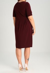 Donna che indossa un vestito bordeaux a lunghezza ginocchio con maniche corte, in piedi su tacchi alti beige, che mostra un tatuaggio sul polpaccio sinistro.