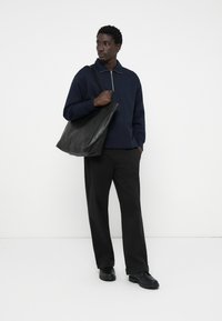 Sudadera azul marino con cremallera y collar, combinada con pantalones de pierna ancha negros y un bolso de cuero negro. La modelo lleva zapatos negros.