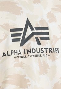 Tan camouflaged t-shirt i bomull, med en framträdande svart logotypdesign med texten "ALPHA INDUSTRIES" och platsinformation.