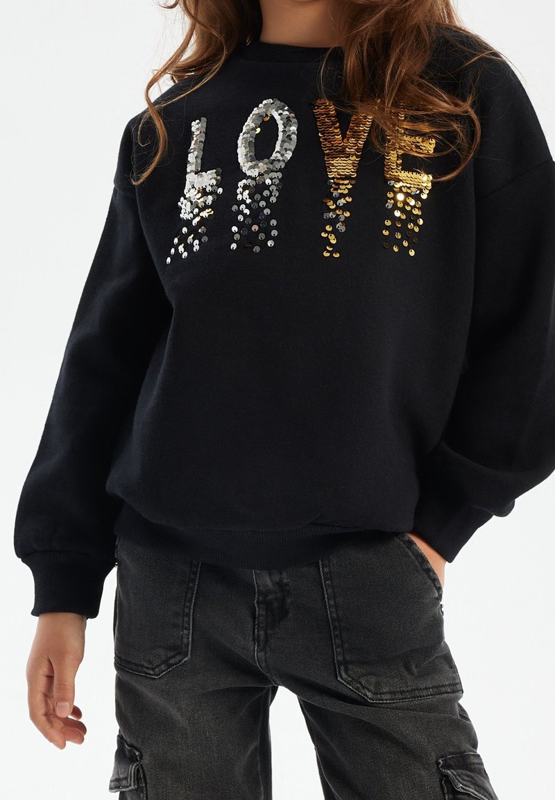 Enfant portant un sweat-shirt noir avec des lettres en sequins dorés et argentés formant "LOVE" et un pantalon cargo gris foncé, main dans la poche.