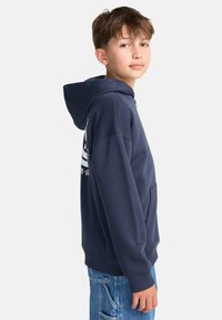 Tmavomodrá mikina s kapucňou, klokaním vreckom, rebrovanými manžetami a bočnými švami. Na zadnej strane má bielu potlač a jemnú textúru.