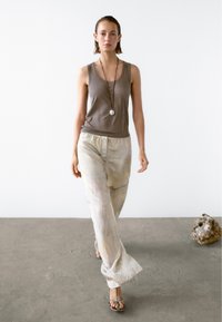 Bruine mouwloze top gecombineerd met lichte, gebloemde wijde broek. Model draagt een lange ketting en sandalen. Neutrale achtergrond.