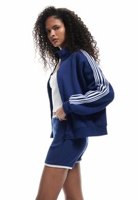 Giacca blu navy con zip e colletto alto, dettagli con tre strisce bianche sulle maniche, abbinata a pantaloni corti blu a coste con orlo a contrasto.