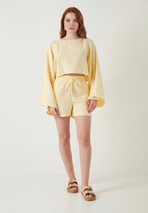 Noor naine seisab, kandes helerohelist pika varrukaga lühikest topi laia varrukaga, sobivaid lühikesi pükse ja beeže sandaale pandlatega.