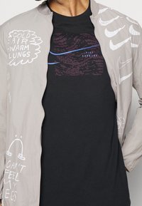 Chaqueta bomber gris claro con texto gráfico blanco y logotipos de Nike, superpuesta a una camiseta negra que presenta un diseño pixelado en tonos morado y azul.