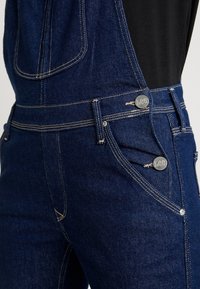 Salopette en denim bleu foncé avec des boutons argentés embossés de "Lee" et des coutures jaunes contrastantes, portée sur une chemise noire.