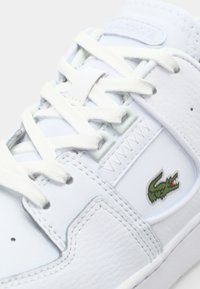 Białe skórzane sneakersy o teksturowanym wykończeniu, z białymi sznurówkami, przeszyciami oraz małym zielonym logo krokodyla Lacoste z boku.