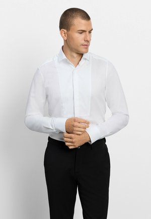 Mann trägt ein weißes Hemd mit Latzfront und schwarze Hose, richtet seinen linken Ärmelbund und schaut nach rechts.
