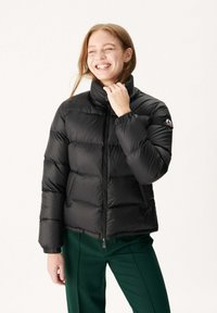 Zwarte pufferjas met een hoge kraag, gestikte stijl en elastische manchetten; gecombineerd met groene broeken, voorzien van een logo op de mouw.