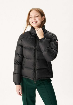 Zwarte pufferjas met een hoge kraag, gestikte stijl en elastische manchetten; gecombineerd met groene broeken, voorzien van een logo op de mouw.