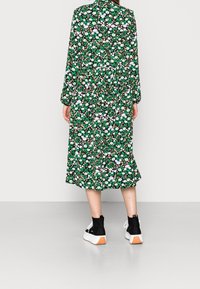 Abito midi floreale verde con maniche lunghe, vita arricciata e un motivo nero, bianco e rosa, abbinato a sneakers nere alla caviglia.