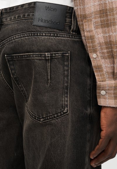 Κοντινό πλάνο τσέπης από σκουρόχρωμο ξεβαμμένο τζιν με δερμάτινη ετικέτα "Won Hundred" σε μαύρο και το χέρι ενός ατόμου που φοράει καφέ πουκάμισο με καρό σχέδιο.