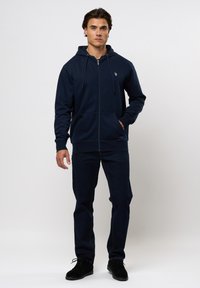 Férfi áll, sötétkék cipzáras kapucnis pulóvert, sötét farmert és fekete cipőt visel, egyszerű világosszürke háttér előtt.