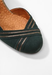 Bottine plate en suède vert avec un bout arrondi, présentant des accents dorés rayés et une semelle intérieure en tan à motifs. Texture douce, design élégant.