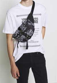 Black and white patterned sling bag with a zip closure, adjustable strap, and two front pockets, worn over a white graphic t-shirt.