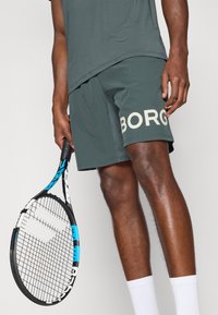 Ciemnozielone szorty sportowe z napisem "BORG" dużymi, jasnymi literami. Czarna rakieta do tenisa z niebieskimi akcentami trzymana w prawej ręce. Widoczne białe skarpetki.