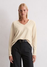 Sweter w kolorze kremowym z dekoltem w kształcie litery V i długimi rękawami, wykonany z miękkiego materiału, połączony z ciemnymi spodniami z wysokim stanem oraz czarnym paskiem ze złotą klamrą.