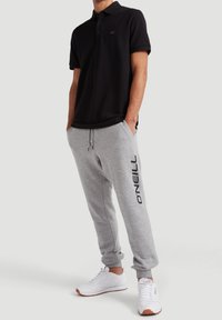 Muž v černém polo tričku s krátkým rukávem, šedých teplácích O’Neill s bočním logem, bílých teniskách, stojící s rukama v kapsách na neutrálním pozadí.