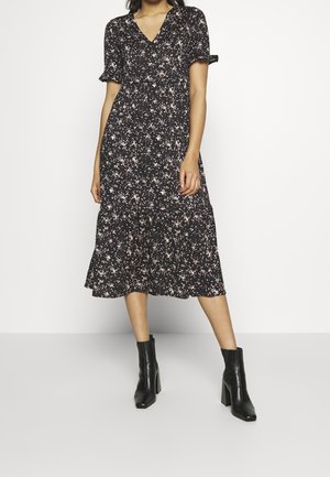 Schwarzes Blumenkleid mit kurzen Ärmeln, V-Ausschnitt und gestuften Rock, das mit einem verstreuten rosa Muster verziert ist. Getragen mit schwarzen, modischen Stiefeletten.