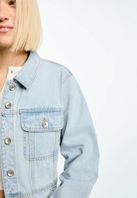 Giubbotto di jeans blu chiaro con una vestibilità strutturata, grande tasca sul petto, bottoni a scatto argentati e aree leggermente sbiadite sul tessuto.