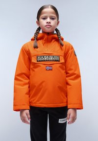 Giacca con cappuccio arancione e tasca frontale, con un patch del logo nero e una bandiera norvegese. Indossata sopra pantaloni neri. Tessuto liscio.