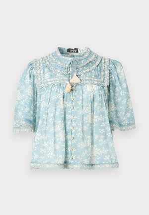 Femme portant un chemisier bleu clair à fleurs avec des manches bouffantes et un short beige, debout avec une main dans la poche et des tongs noires.