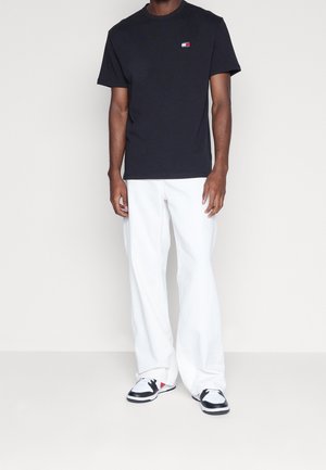 Musta lyhythihainen puuvillainen T-paita, jossa on pyöreä pääntie ja pieni monivärinen logo. Yhdistetty valkoisiin leveälahkeisiin housuihin ja mustiin lenkkareihin.