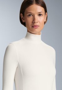 Bijela majica s lažnim rolkama napravljena od glatkog, rastezljivog materijala. Prilagođen dizajn s dugim rukavima, bez vidljivih uzoraka ili metalnih detalja.