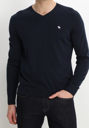 Homme portant un pull col en V bleu marine avec un petit logo élan blanc sur la poitrine, associé à un jean foncé, main dans la poche.