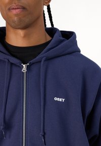 Navy blue zip-up hoodie made of soft fabric, featuring a drawstring hood, white "OBEY" text on the chest, and silver zipper hardware.