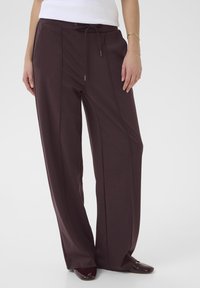 Pantaloni ampi di colore marrone scuro realizzati in tessuto morbido, con vita regolabile tramite cordino, pieghe frontali e una finitura liscia.