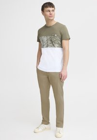 Olive green and white color-blocked T-shirt with a textured upper section, paired with beige trousers and cream sneakers.