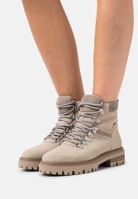 Beige Wildleder-Ankleboots mit einem strukturierten Kragen, metallischen Ösen und stützender Schnürung. Massive Gummisohle mit tiefen Profilrillen für bessere Traktion.