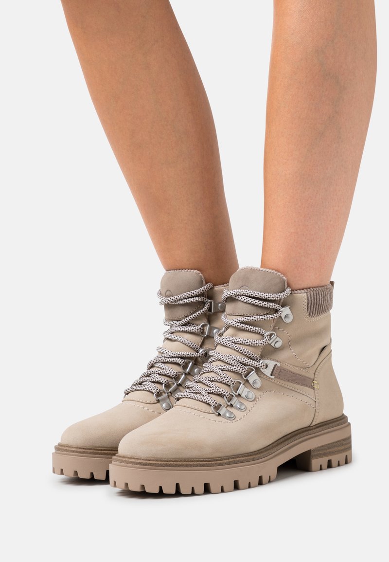 Beige Wildleder-Ankleboots mit einem strukturierten Kragen, metallischen Ösen und stützender Schnürung. Massive Gummisohle mit tiefen Profilrillen für bessere Traktion.