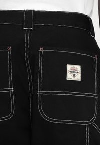 Black work pants with white stitching, featuring two large back pockets, a tag with "Worn Wear Ice Cream," and a durable fabric texture.