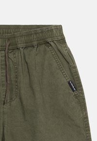 Λαστιχένια βερμούδα σε ελιά πράσινη με τσέπες εμπρός και σκοτεινό κορδόνι, με μια μικρή μαύρη ετικέτα της μάρκας στη ραφή της τσέπης.