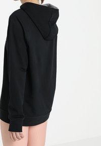 Svart hoodie med huva, långa ärmar och en avslappnad passform. Materialblandning i bomull, synlig sömnad och ribbade ärmslut.