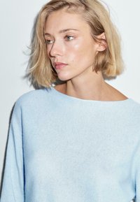 Giovane donna dai capelli biondi corti che indossa un maglione blu chiaro, che guarda di lato su uno sfondo bianco uniforme.
