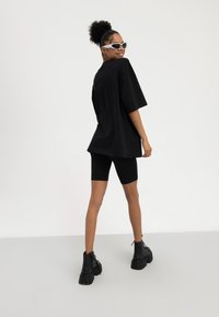 Kvinne med krøllete hår i høy knute har på seg en oversized svart t-skjorte, svarte biker-shorts, kraftige svarte støvler og hvite solbriller, smilende bakover.