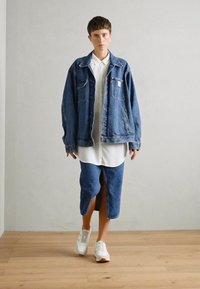 Jeansjacke in heller Waschung mit Fronttaschen, kombiniert mit einem weißen Button-Down-Hemd und einem knielangen Jeansrock mit Schlitz vorne. Weiße Turnschuhe.