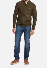Uomo che indossa un maglione olive verde con zip, jeans blu e sneakers in suede marrone, in piedi davanti a uno sfondo neutro.