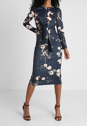 Mujer con vestido floral azul marino de manga larga y largo hasta la rodilla, con detalle de lazo en el frente y tacones burdeos con tiras en el tobillo, posando frente a un fondo gris.