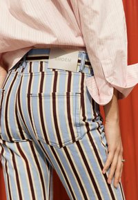 Persona vistiendo pantalones vaqueros con rayas verticales en azul, blanco, marrón y amarillo con una etiqueta BHOËM, combinados con una camisa a rayas rosa pálido sobre un fondo rojo.