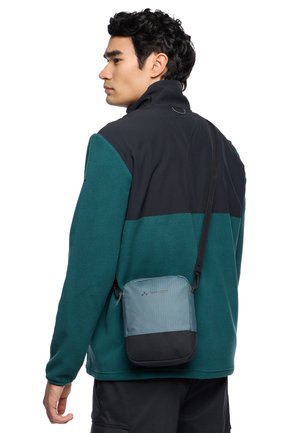 Sac en bandoulière gris et noir avec fermeture éclair, surface texturée, sangle ajustable et détail de logo, porté sur une veste polaire turquoise et noire.