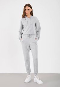 Vrouw staat met handen in haar zakken, gekleed in een lichtgrijze hoodie, bijpassende joggingbroek en witte sneakers op een effen achtergrond.