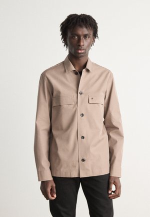 Camisa de botones marrón claro con dos bolsillos en el pecho, botones negros y un cuello, llevada sobre pantalones negros. Textura de tela suave.