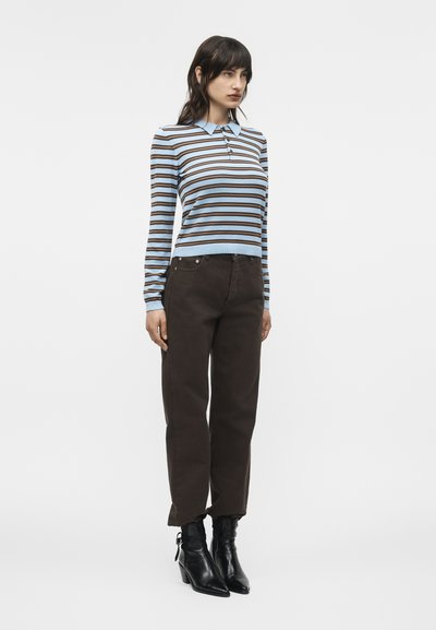 Femme portant un polo à manches longues rayé bleu et marron, un pantalon marron foncé et des bottines noires, debout devant un fond blanc.